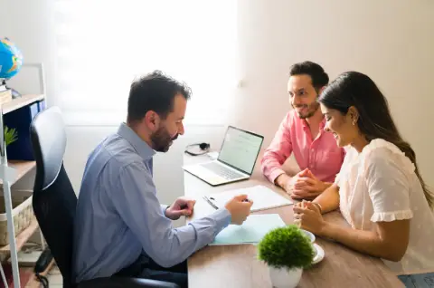 A man in a blue shirt discusses paperwork with a couple at a desk, with a laptop and a small plant present